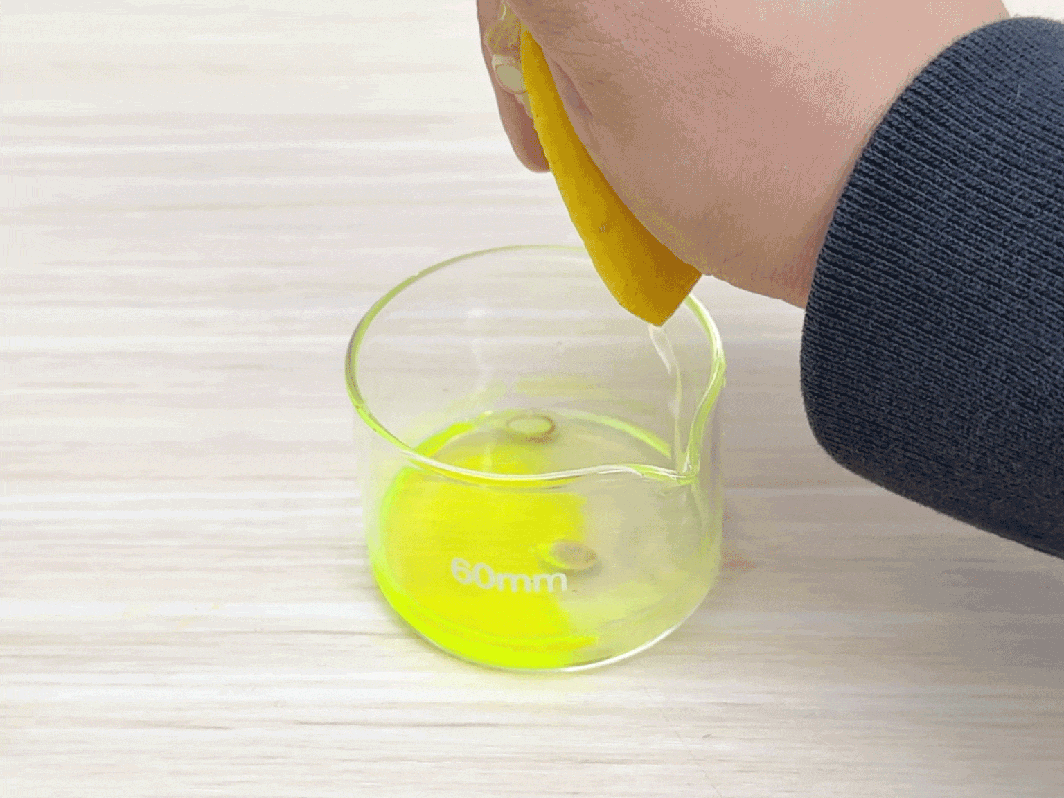 A hand squeezes a cut lemon half over a small, clear glass beaker containing a bright, fluorescent yellow liquid. The beaker sits on a light woodgrain surface. Drops of lemon juice are visible falling from the lemon into the beaker. This image suggests an experiment or demonstration where lemon juice is being added to a pre-existing solution, likely to observe a chemical reaction or color change.