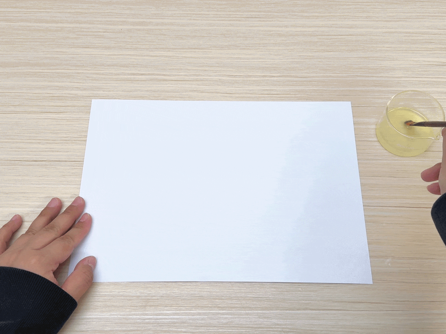 A blank white sheet of paper lies on a light woodgrain table. A hand is partially visible, dipping a small paintbrush or similar tool into a clear container filled with a pale yellow liquid. Another hand rests on the edge of the paper, seemingly holding it in place. This image suggests an art project or experiment involving applying the liquid to the paper, likely to create a design or observe a reaction.