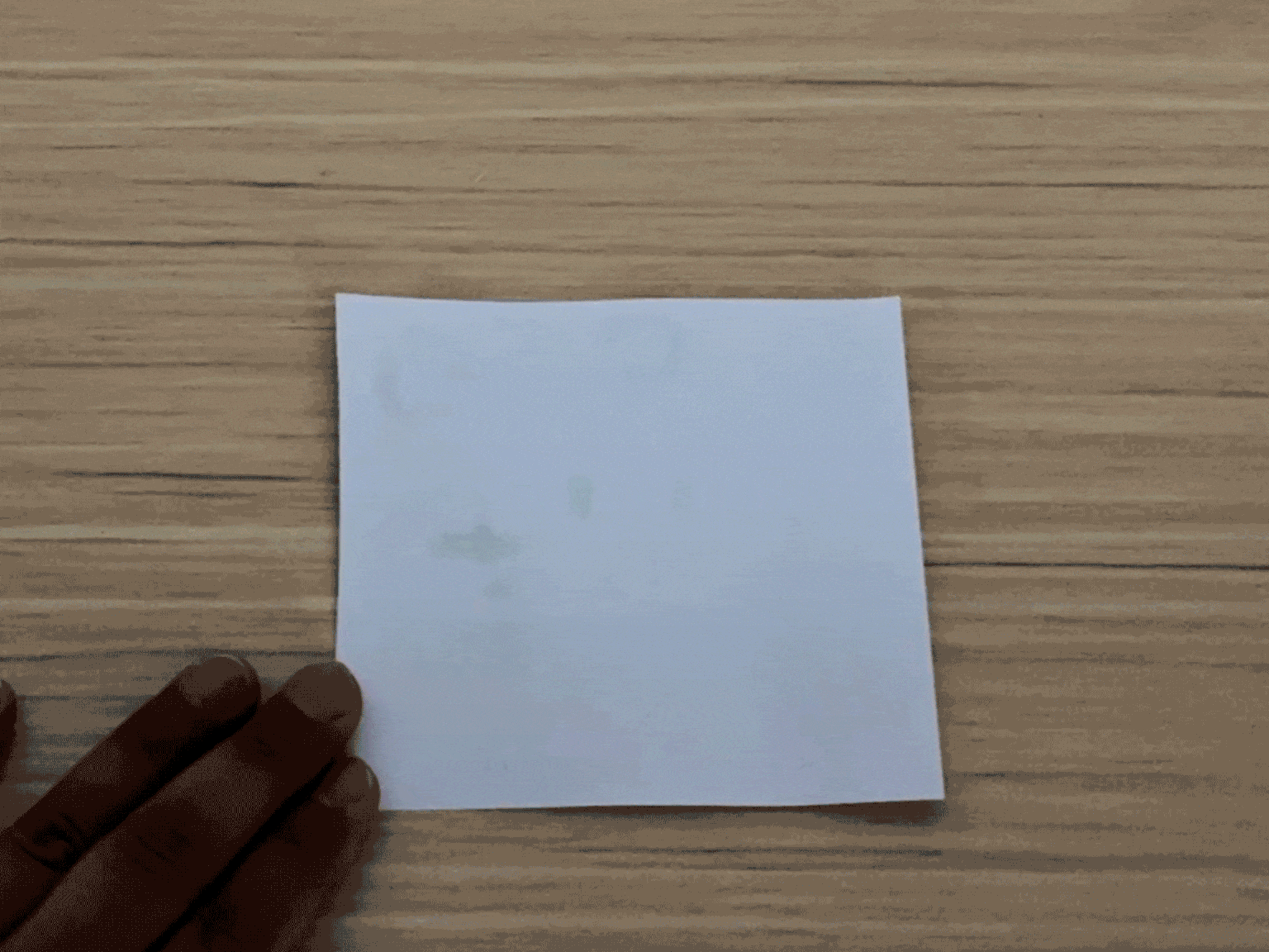 A hand partially visible in the bottom left corner holds a piece of white paper against a light woodgrain table. The paper has a few faint, light-colored markings on it, possibly from a liquid or light heat exposure. The focus is on the paper and its subtle markings, suggesting the result of an experiment or a creative process.