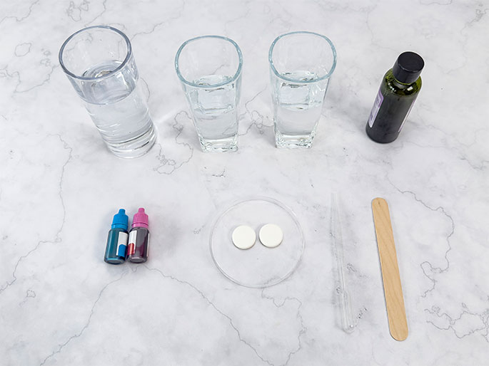 An assortment of craft supplies is arranged on a light wood table. These include four small, clear glass jars (two pairs placed side-by-side), a white coffee filter, a group of colourful markers in shades of purple, pink, red, and blue, a pair of scissors, and a black pipe cleaner. The items appear to be laid out in preparation for a craft project.