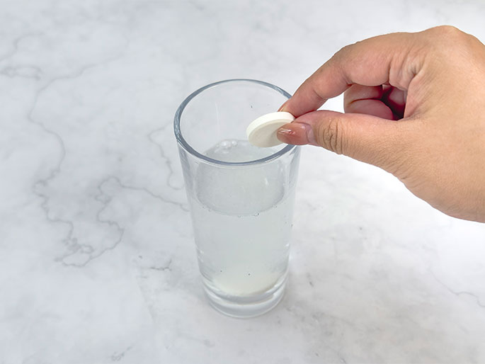 Two hands gently flatten a round white coffee filter on a light wooden surface. The filter's pleated edges are visible, and the hands hold it in place, presumably in preparation for a craft or experiment.