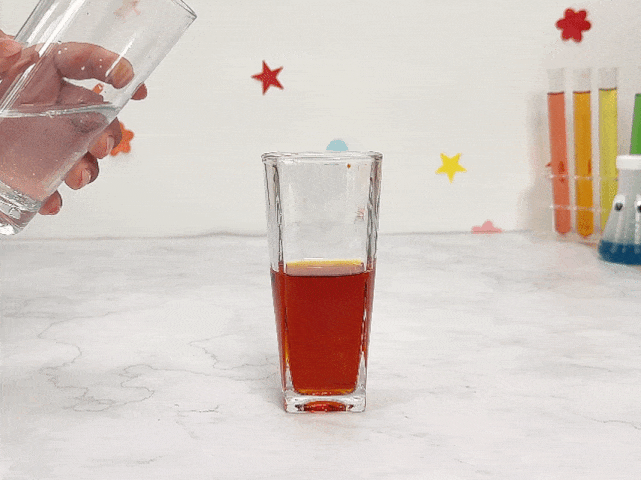 A hand pours clear water from a glass into a tall, square glass containing a small amount of orange liquid. As the water mixes with the orange liquid, it creates a swirling, cloudy effect, gradually diluting the colour. The glass sits on a marble surface. In the background, colourful star and flower stickers decorate the wall, and a rack of test tubes with various coloured liquids is visible. The video demonstrates a simple mixing experiment, possibly exploring colour dilution or density differences between liquids.
