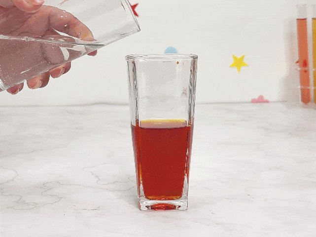 Clear water is poured from a glass into a tall, square glass containing a small amount of red liquid. The water mixes with the red liquid, creating a swirling, cloudy effect as it dilutes the colour. The glass sits on a marble surface. Colourful star and flower stickers decorate the wall in the background. A rack of test tubes containing various coloured liquids is also visible. This video likely demonstrates a mixing experiment, possibly exploring colour dilution or the differences in density between the liquids.