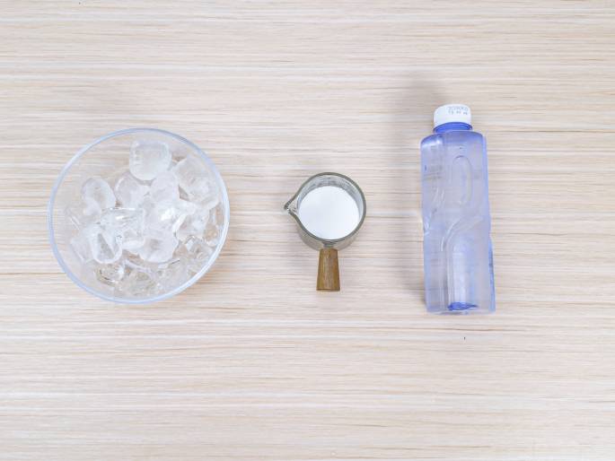 A top-down view of ingredients arranged on a light wood surface. A clear bowl of ice cubes is on the left, a small glass pitcher with a wooden handle containing a white liquid (presumably milk or cream) is in the center, and a plastic bottle of water is on the right. These items appear ready for preparing a cold beverage.