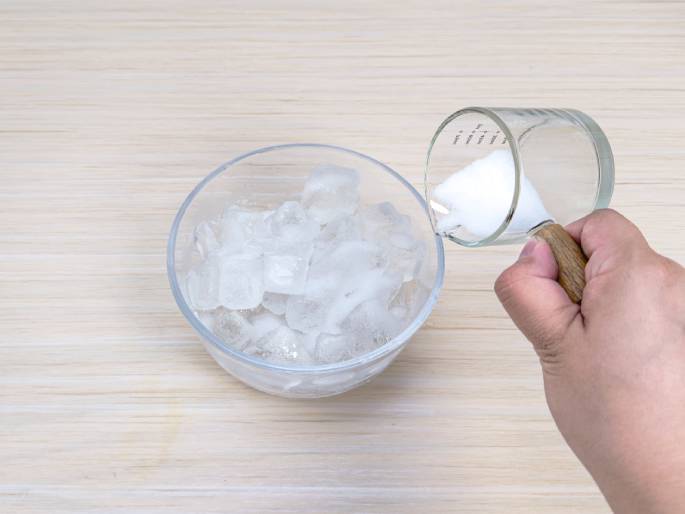 A hand pours a white powder, likely baking soda or a similar substance, from a clear glass measuring cup with a wooden handle into a clear bowl filled with ice cubes. The process is taking place on a light wooden table or countertop. This image suggests a step in a science experiment or the preparation of a cold beverage or other recipe.
