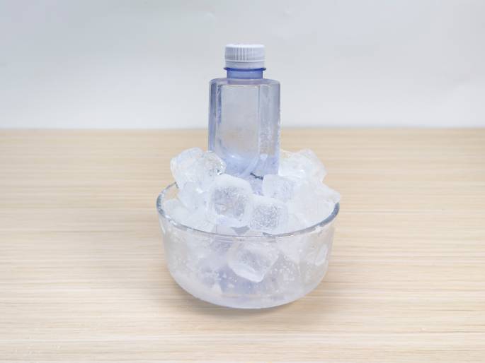 A small, clear plastic bottle of water rests on a mound of ice cubes in a clear glass bowl. The bowl and bottle are on a light wood surface. Condensation is visible on the bottle, indicating it is cold. This image likely depicts a method for quickly chilling a bottle of water.