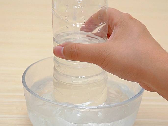 A hand holds a clear plastic bottle, partially filled with a clear liquid, and places it into a clear glass bowl filled with ice water and ice cubes. The bottle is being submerged into the icy water to cool its contents. The background is a light wooden surface.