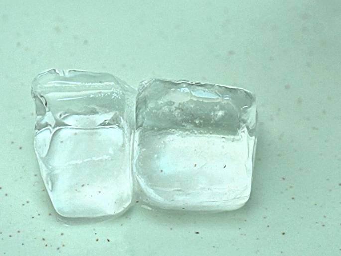 Two ice cubes sit on a pale green surface. They are slightly melting, as evidenced by the glistening surfaces. The surface underneath appears to have small, dark speckles.