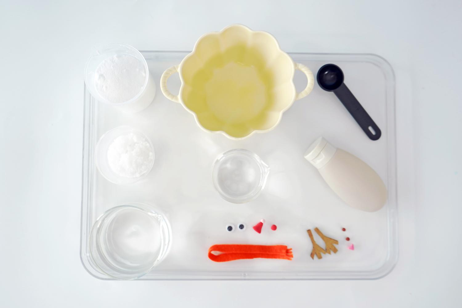 A clear plastic tray neatly organises the components for a melting fizzy snowman experiment. Items include two small containers of white powder (likely bicarbonate of soda), a small bowl of clear liquid (vinegar), a larger pale yellow bowl (for mixing), a small measuring spoon, a bottle of what seems to be cream or a similar liquid, and small decorative elements such as googly eyes, a red nose, and twig-like arms for the snowman. The tray sits on a plain white surface. The overall arrangement is clean and clearly indicates preparation for a fun, science-based activity.