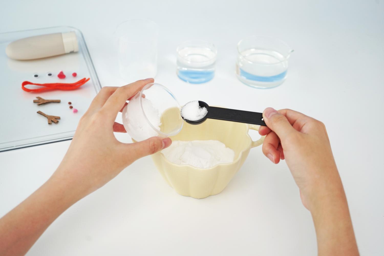 Two hands are adding bicarbonate of soda to a pale yellow bowl using a small black measuring spoon. A small glass bowl containing more bicarbonate of soda is held in one hand, while the other hand holds the spoon, carefully transferring the powder. In the background, a clear tray holds additional ingredients and snowman decorations, including two small bowls of water, a bottle of cream, and small craft items such as eyes and a nose. The scene is set on a plain white surface. The image clearly shows a step in the preparation for a melting fizzy snowman experiment.