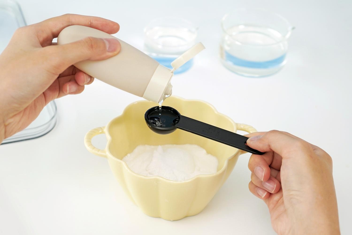 A person is adding vinegar to bicarbonate of soda in a pale yellow bowl. Vinegar is being poured from a small beige bottle into a black measuring spoon, which is then being used to add the vinegar to the bicarbonate of soda. In the background, there are additional containers of water and snowman decorations, suggesting the preparation for a melting fizzy snowman experiment. The setting is a clean, white surface. The image focuses on the careful addition of vinegar, a crucial step in the experiment.