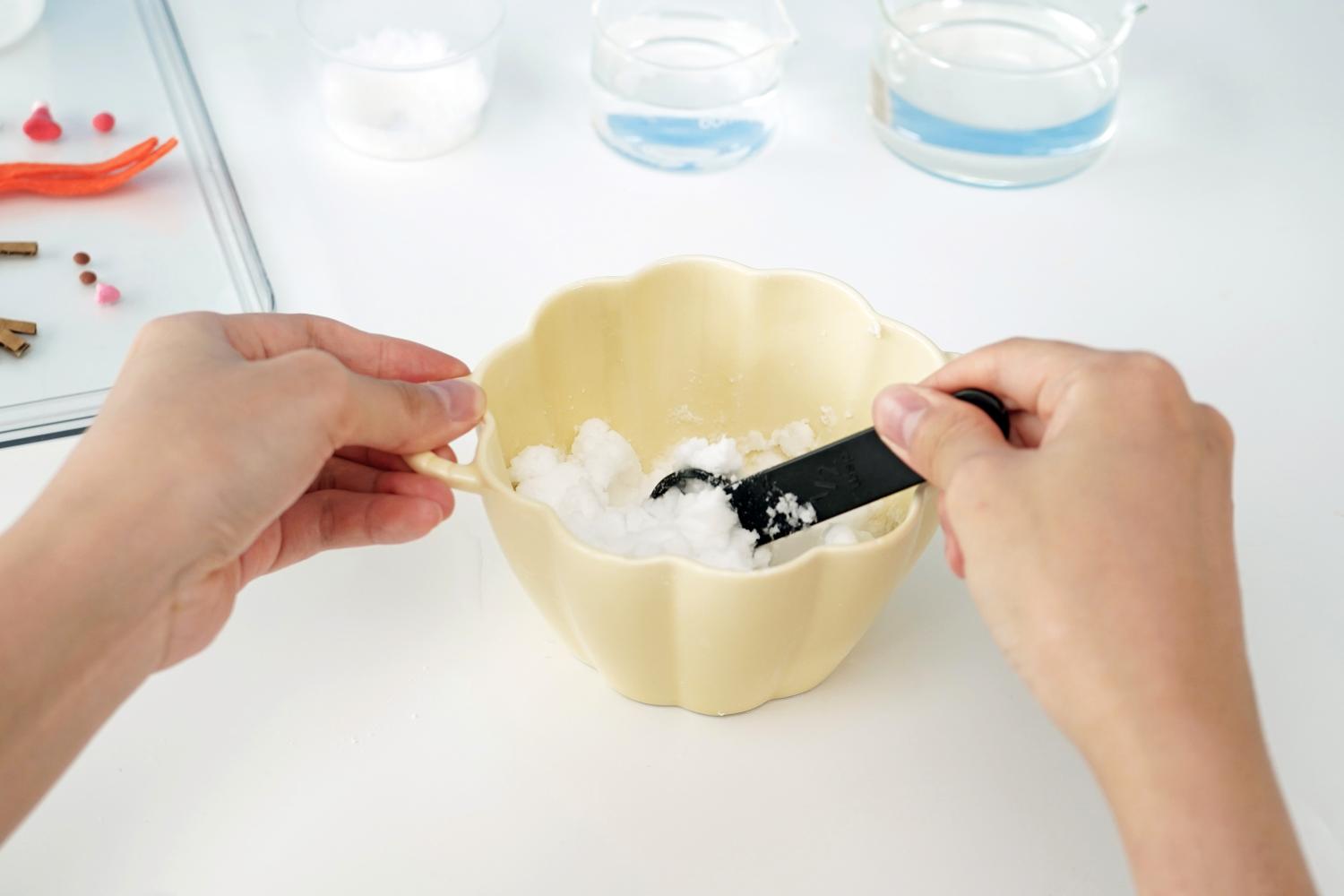 Two hands are adding bicarbonate of soda to a pale yellow bowl using a small black measuring spoon. The bicarbonate of soda is being transferred from a small glass bowl into the larger one. In the slightly blurred background, a clear tray with snowman-making decorations and small containers of water are visible. The entire scene is on a plain white surface. The image shows a step in the preparation for a melting fizzy snowman experiment, focusing on the careful measuring and transfer of the bicarbonate of soda.