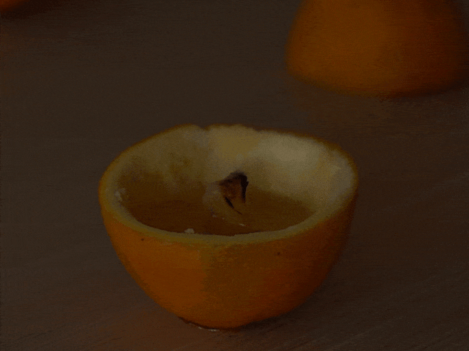 A small, makeshift candle is created using an orange peel as a container. The orange has been cut in half, and the pulp removed, leaving the rind. A small piece of wick or string sits in a pool of orange oil or juice inside the bottom half of the rind. A lit match or lighter ignites the wick, creating a small flame. The top half of the orange rind is then placed back on top, partially covering the flame and creating a small, enclosed orange candle. The video demonstrates a simple DIY project for creating a candle using an orange.