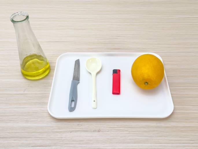 Arranged on a light woodgrain table are several items, suggesting preparation for an experiment or demonstration. A clear glass bottle containing a yellow liquid, possibly oil, sits to the left. A white rectangular tray holds a small knife with a gray handle, a white plastic spoon, a red lighter, and a whole, ripe orange. The items are neatly placed on the tray, implying a deliberate setup for a specific purpose.
