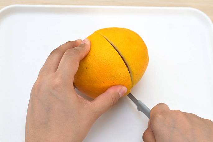 Two hands are shown in the process of cutting an orange on a white cutting board or tray. One hand holds the orange steady while the other hand uses a small knife with a gray handle to slice into the fruit. The orange is being cut lengthwise, and the cut is partially visible. The image suggests a simple food preparation task.