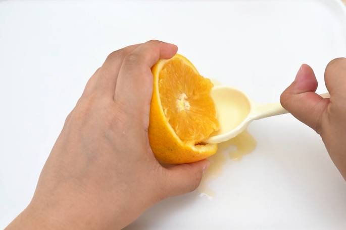 Hands are using a white plastic spoon to extract juice from a halved orange on a white surface. One hand holds the orange half while the other uses the spoon to press against the fruit's flesh, releasing juice. A small amount of orange juice is visible on the white surface.