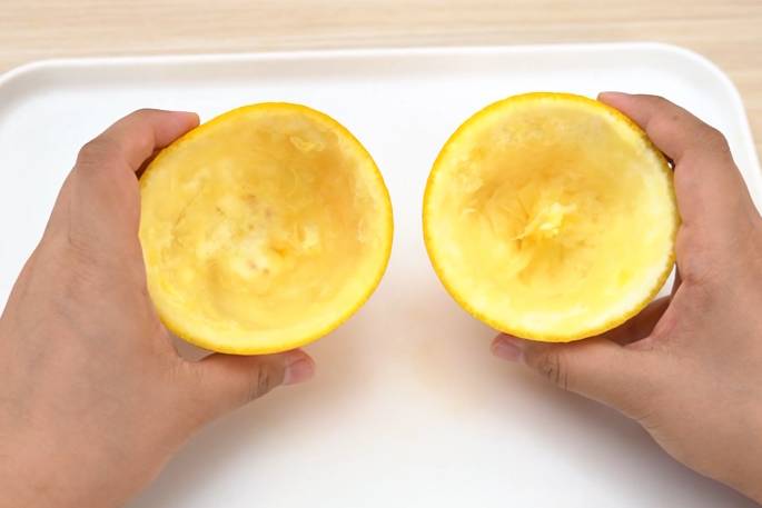 Two hands hold the two hollowed-out halves of an orange over a white surface. The orange rinds are mostly empty of pulp and juice, suggesting they have been thoroughly squeezed or scooped out. The image focuses on the inside of the orange peels.
