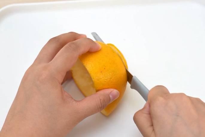 Two hands are shown carefully peeling an orange with a small knife on a white surface. One hand holds the orange while the other hand uses the knife to remove the peel. A section of the peel is being lifted away from the fruit. The focus is on the peeling process.