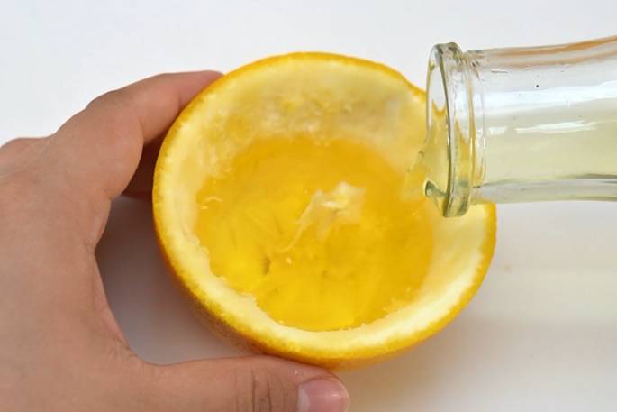 A hand holds a hollowed-out orange half while a clear glass bottle pours a yellow liquid, presumably oil, into it. The oil is flowing into the orange peel, which acts as a makeshift container. The process suggests an unusual or experimental use of the orange peel.