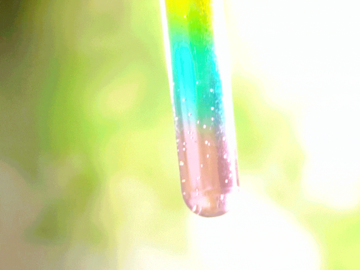 A test tube, filled with a layered rainbow of liquids, is held up against a blurred green background. The colours transition smoothly from yellow at the top, through green and blue, to pink at the bottom. Tiny bubbles are visible within the liquids, particularly in the lower sections, suggesting a recent mixing or chemical reaction. The bright colours and the presence of bubbles create a visually dynamic and engaging image.