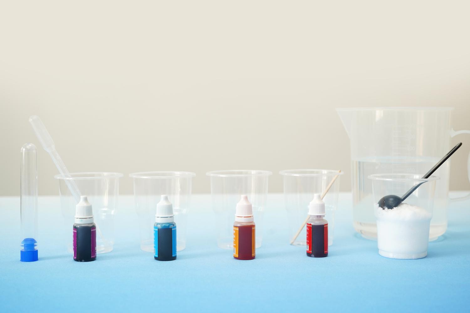 A light blue surface displays the meticulously arranged components for a Rainbow in a Tube experiment. Five small bottles of coloured liquid, exhibiting varying shades of red, orange, yellow, blue, and purple, are centrally positioned. These are flanked by clear plastic beakers, a larger clear plastic jug of water, a small container of white powder (likely sugar or salt), a measuring spoon, a pipette, and a small stirring stick. The setup is neat and organised, ready for the layering process to create the colourful density column.