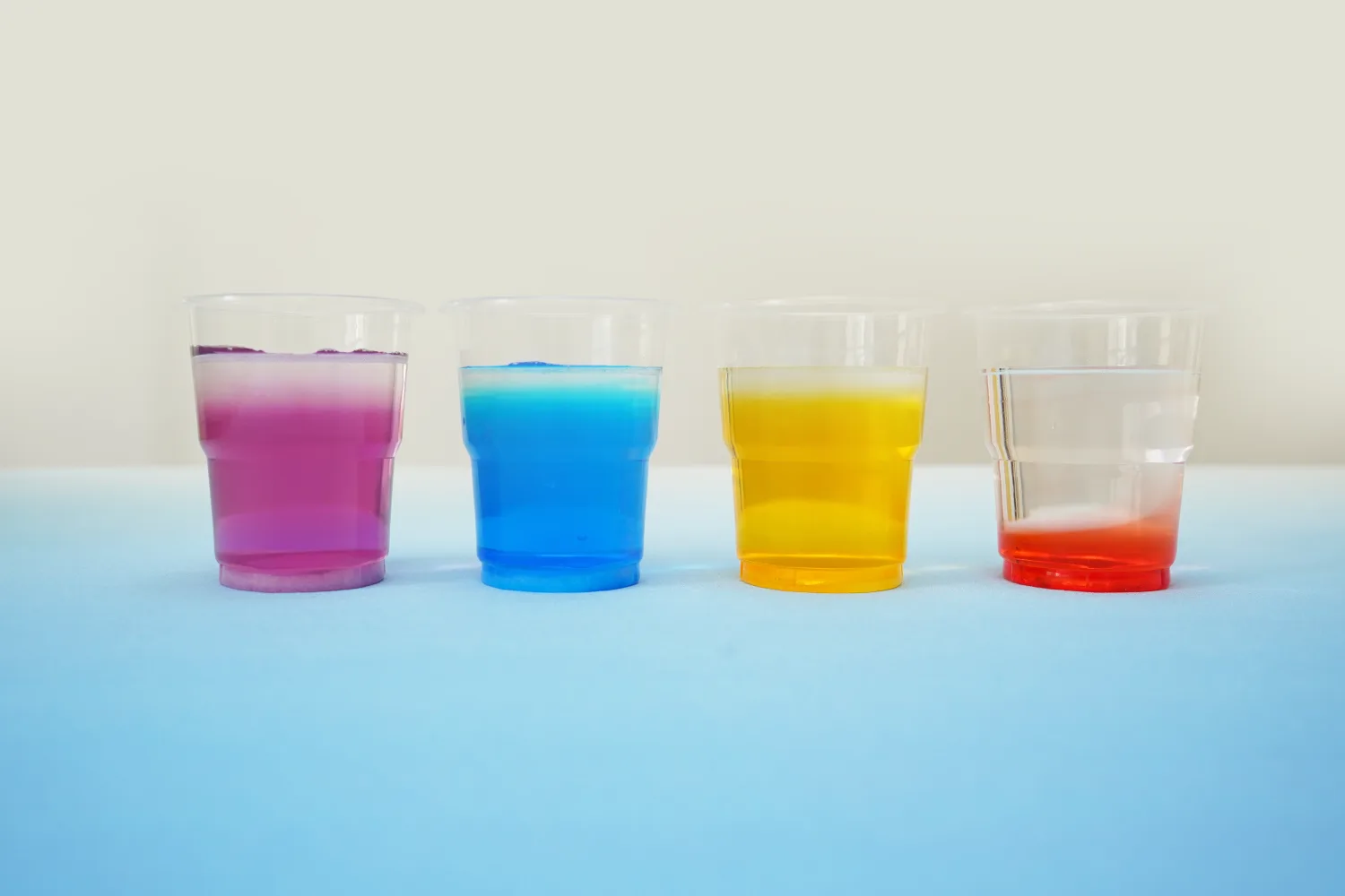 Four clear plastic cups, arranged in a row on a pale blue surface, contain layered liquids creating a miniature rainbow effect. From left to right, the cups show layers of purple, blue, yellow, and red liquids, with a thin white layer visible at the top of each. The colours are vibrant and distinct, clearly demonstrating the principle of liquid density. The background is a plain, off-white wall, drawing attention to the colourful cups. The overall impression is of a successful and visually appealing science experiment.
