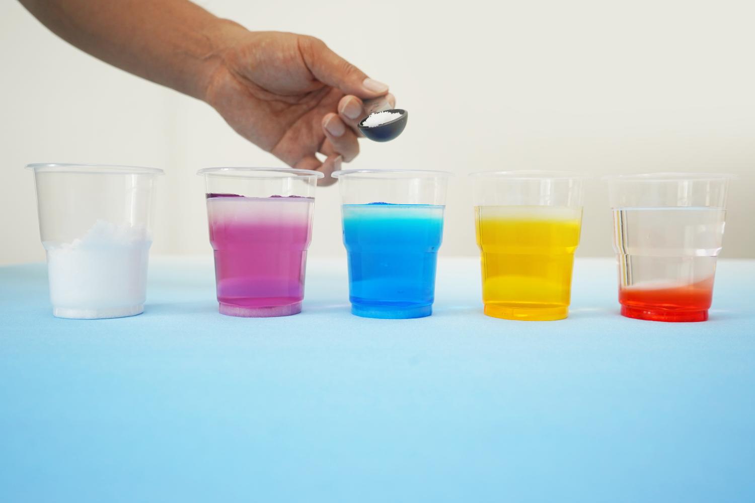 A hand is adding a small amount of white powder, likely sugar or salt, to a clear plastic cup containing a layered liquid. The cup is one of four arranged in a row on a light blue surface, each cup containing a different coloured liquid, creating a rainbow effect. The colours are vibrant and distinct, showing purple, blue, yellow, and red. The background is a plain, off-white wall. The image shows a step in a density experiment, focusing on the addition of a substance to alter the density of one of the layers.