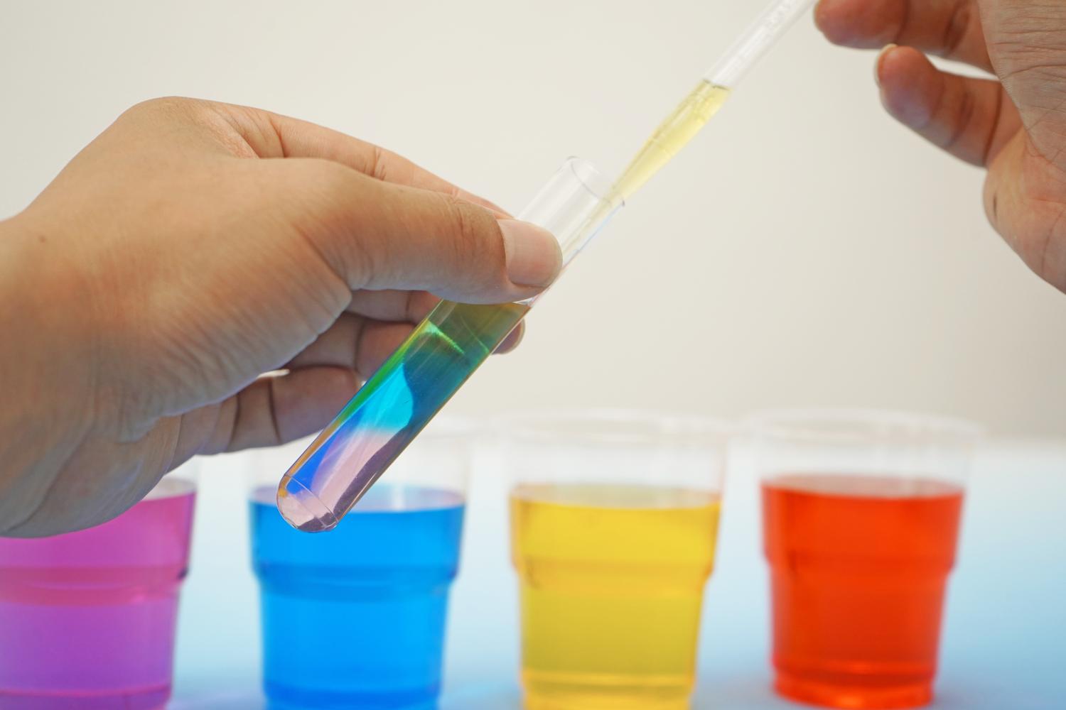 A hand gently stirs a purple liquid in a clear plastic cup using a small wooden stick. This cup is one of four arranged on a light blue surface; the others contain blue, yellow, and red liquids respectively, creating a rainbow effect. The background is a plain, off-white wall. The image shows a step in a density experiment, illustrating the careful mixing of one of the coloured layers to ensure a smooth, even distribution of colour.
