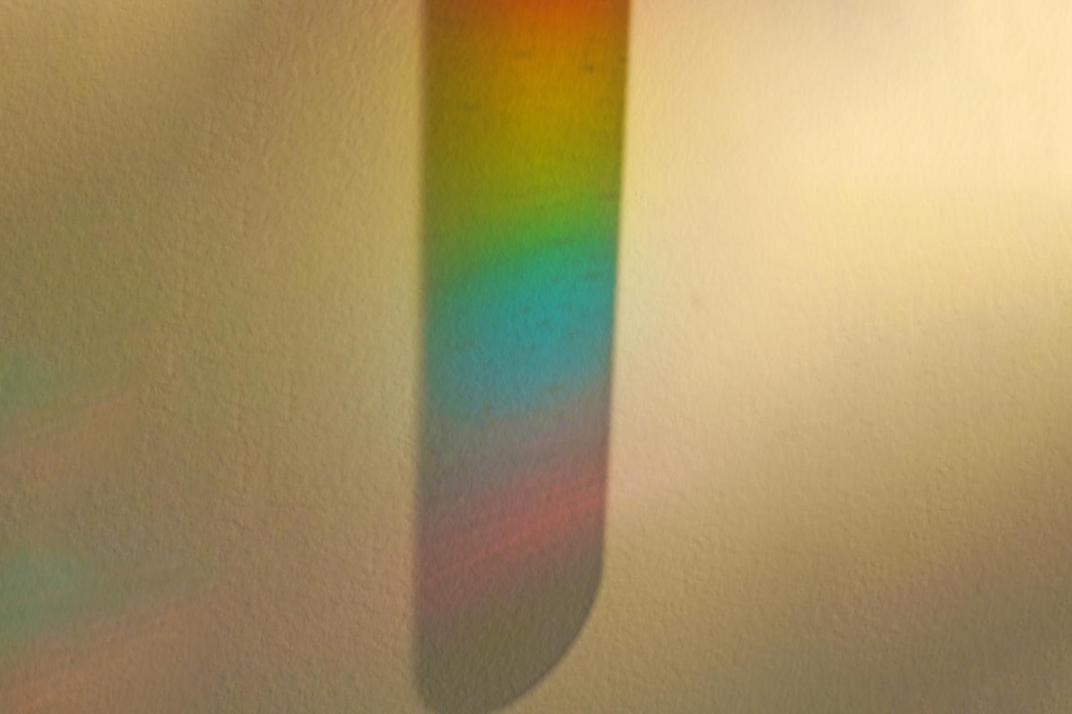 The image shows a blurred, slightly out-of-focus projection of a rainbow onto a textured, off-white surface. The colours, though indistinct due to the blur, appear to be layered vertically, suggesting a projection from a cylindrical object containing layered liquids, similar to a rainbow in a tube experiment. The background is a muted, neutral tone, allowing the projected rainbow to be the focal point. The overall effect is soft and ethereal, hinting at the underlying science experiment.