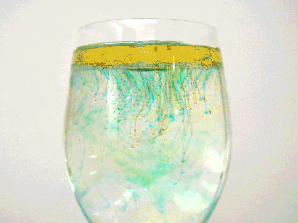 A clear wine glass is partially filled with a layered liquid. A distinct layer of yellow oil or a similar substance floats on top, while beneath it, wispy tendrils of green and blue food colouring or dye create swirling patterns in the water. Tiny bubbles are also visible within the water, adding to the visual effect. This arrangement likely demonstrates the interaction of oil and water, as well as the diffusion of colour in a liquid medium.