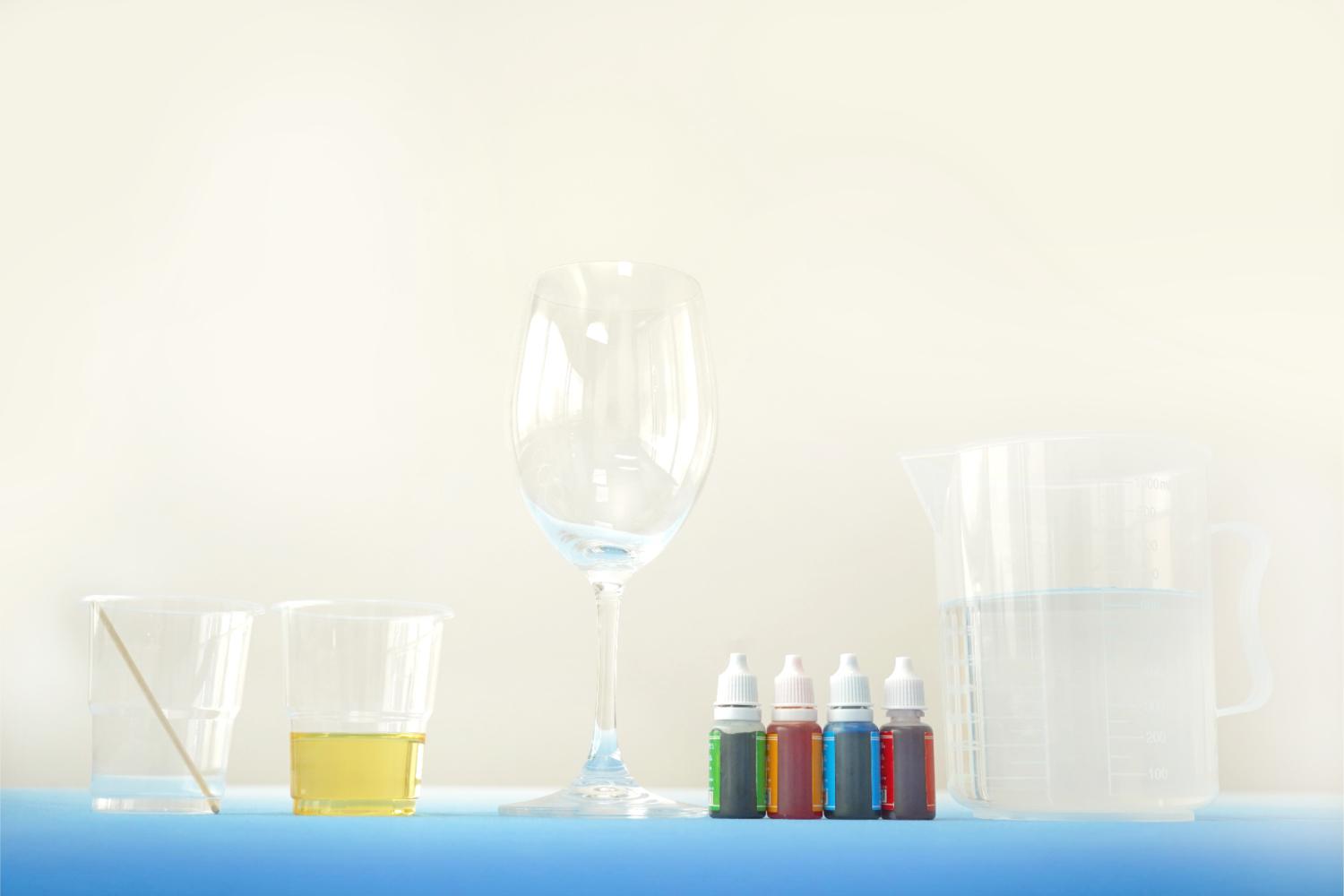 On a light blue surface, against a pale background, the equipment for a Rainbow Rain in a Glass experiment is neatly arranged. A wine glass sits centrally, flanked by two clear plastic cups, one containing water and a small wooden stirrer, the other holding a yellowish liquid (likely oil). Four small bottles of coloured liquid, in red, orange, yellow and blue, are lined up in front of the wine glass. A larger clear plastic jug of water completes the setup. The overall arrangement is clean, organised, and clearly shows the preparation for a science experiment.