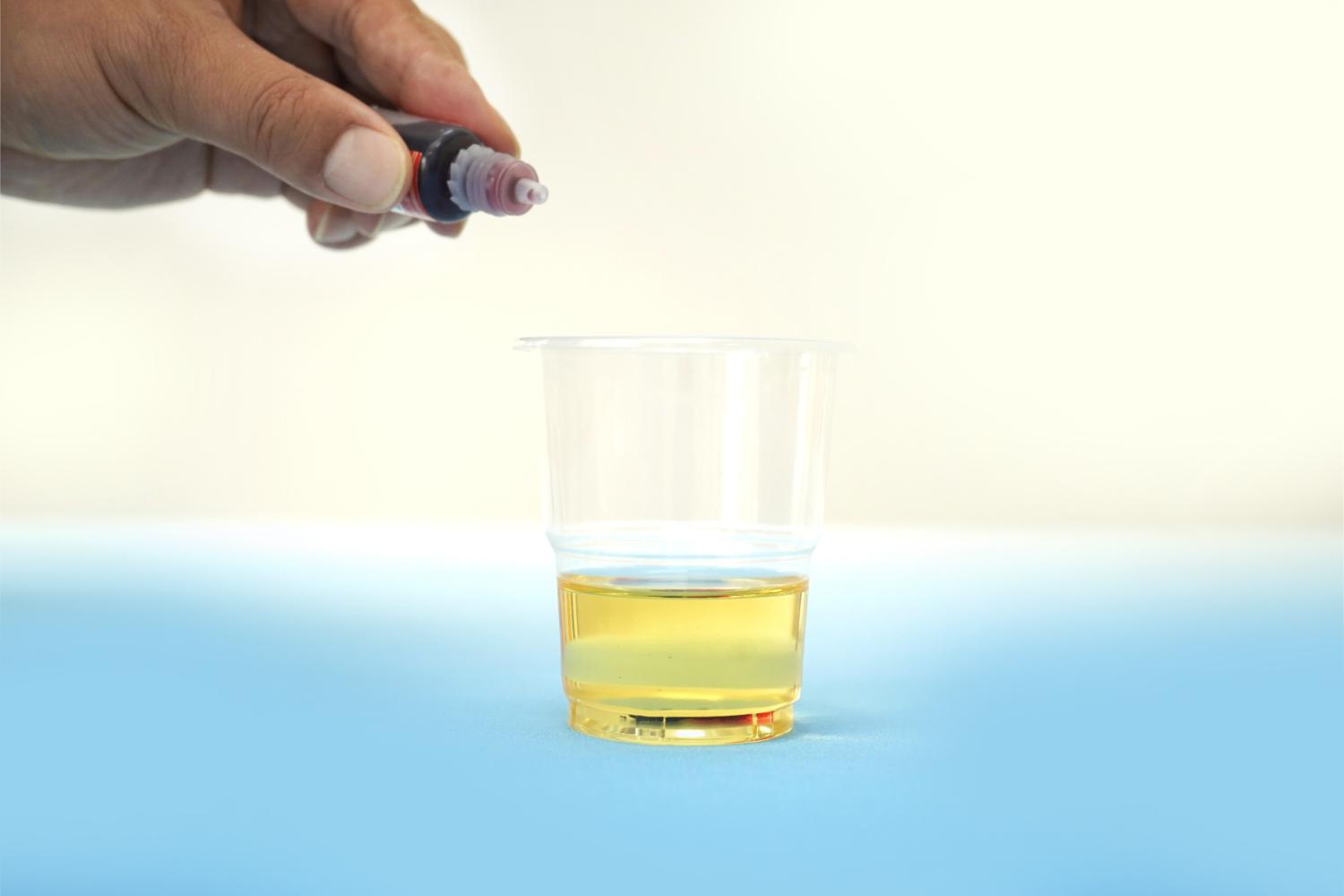 A hand adds coloured liquid from a small bottle to a clear plastic cup containing a yellowish liquid, on a pale blue surface.