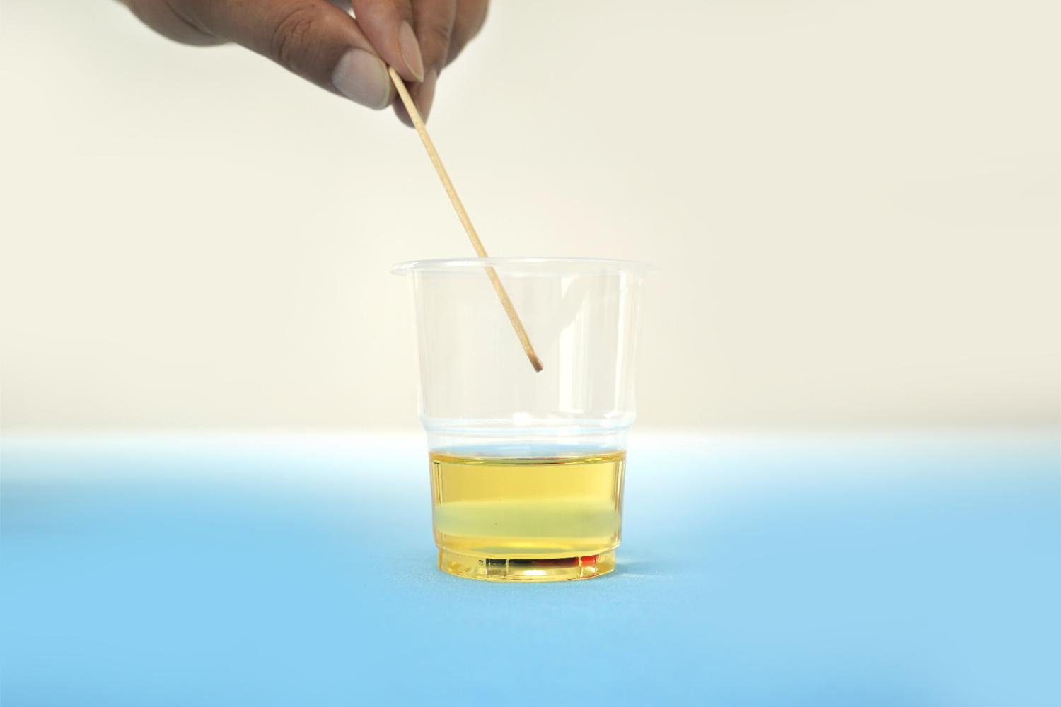 A hand gently stirs a yellowish liquid in a clear plastic cup using a small wooden cocktail stick, against a pale blue background.