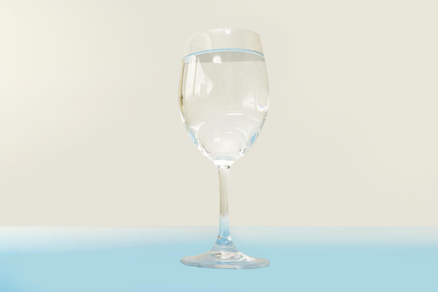 A clear wine glass, partially filled with water, stands on a pale blue surface against a cream-coloured background.