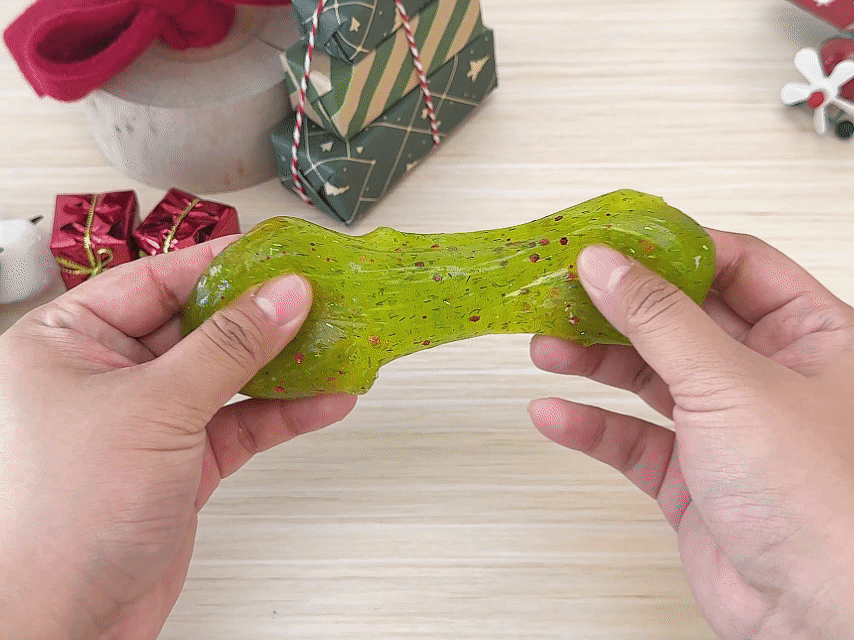 Two hands manipulate a bright, lime-green slime. The slime is translucent and filled with small, sparkly red and green glitter or confetti. The hands stretch and pull the slime, demonstrating its viscous and stretchy texture. In the background, there are Christmas-themed decorations, including small wrapped presents, a miniature Christmas tree made of stacked boxes, and a Santa Claus hat. The video showcases the playful qualities of festive, glittery slime against a Christmas backdrop.