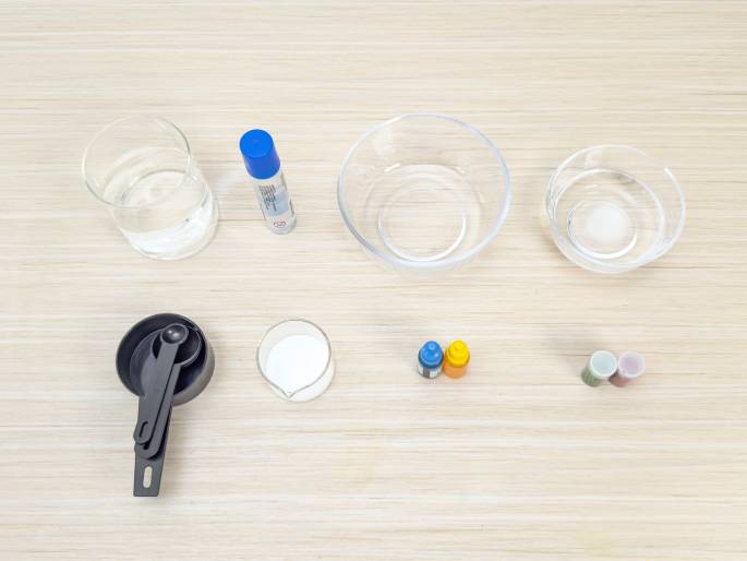 An overhead flatlay shot displays various ingredients and tools laid out on a light wood-grained surface, seemingly for a craft or science project. Items include clear glass bowls and cups containing water and a white substance, a set of black measuring spoons, small bottles of blue and yellow coloring, a glue stick, and small containers of glitter or colored powder. The arrangement suggests preparation for a specific activity.