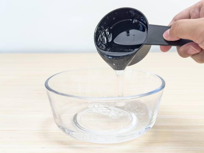 A hand pours a clear, viscous liquid from a black measuring cup into a clear glass bowl. The liquid flows in a steady stream from the cup to the bowl, which is placed on a light wooden surface. The scene suggests a step in a recipe or experiment where a precise amount of liquid is being added.
