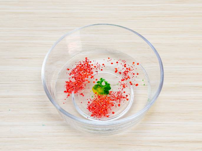 A clear glass bowl sits on a light wooden surface, containing a mixture of water, red glitter, and a small drop of green and yellow coloring. The glitter is dispersed throughout the water, while the coloring remains concentrated in a small area. This setup suggests an experiment or craft activity involving color mixing or glitter effects.