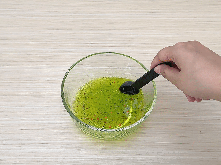 A hand uses a small black measuring spoon to stir a mixture in a clear glass bowl. The mixture is a viscous, yellowish-green liquid with red glitter dispersed throughout. The bowl sits on a light wood-grained surface. The action suggests the combining of ingredients or the thickening of a substance, likely for a craft or science project.