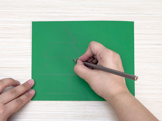 A hand draws a Christmas tree shape on a piece of green card with a pencil on a light wood-grain surface.