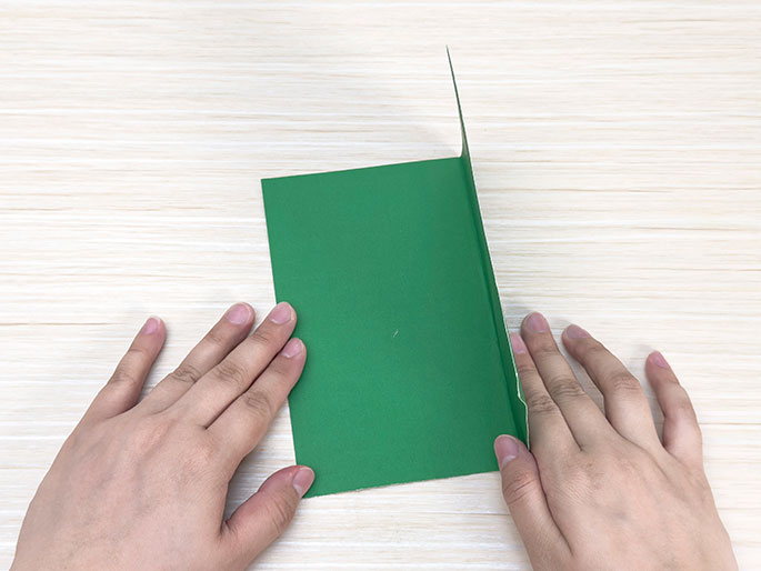Two hands fold a piece of green card in half on a light wood-grain surface.