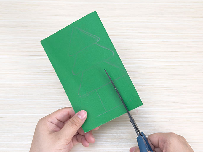 Two hands use scissors to cut along a pencil line of a Christmas tree shape drawn on a piece of green card on a light wood-grain surface.