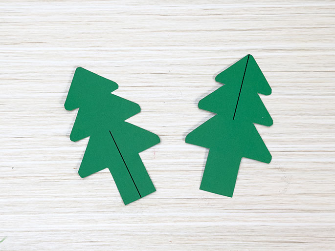 Two identical Christmas tree shapes cut from green card, each with a vertical line drawn down the centre, lie on a light wood-grain surface.