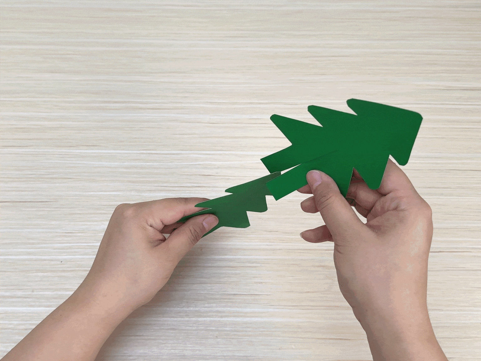 Two hands slot two identical Christmas tree shapes cut from green card together on a light wood-grain surface.