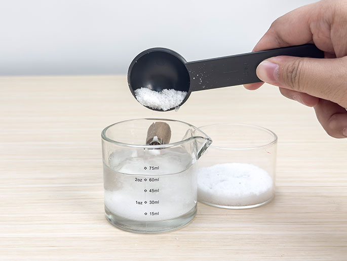 A hand pours white crystals from a black measuring spoon into a glass beaker of water, next to a small glass dish of the same white crystals, on a light wood-grain surface.