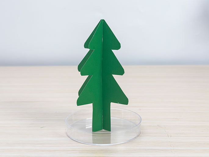 A small, green, cardboard Christmas tree stands in a clear, shallow dish on a light wood-grain surface.