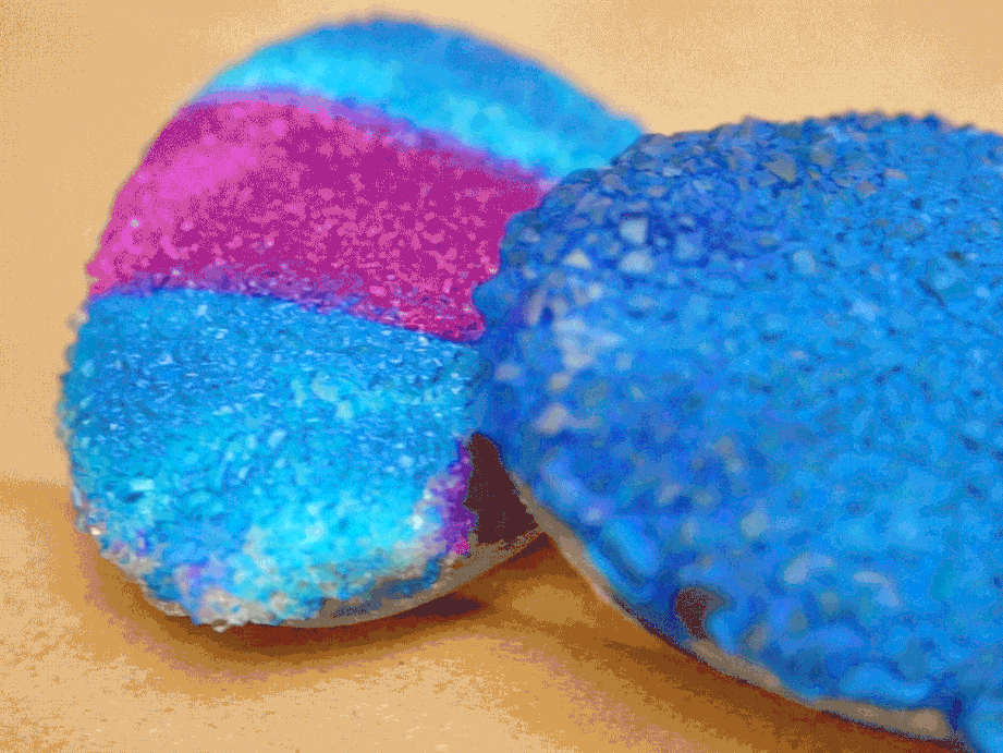 Two small, round objects, possibly bath bombs or cookies, are covered in coarse, brightly coloured sugar crystals, one with distinct blue and pink bands and the other entirely blue.