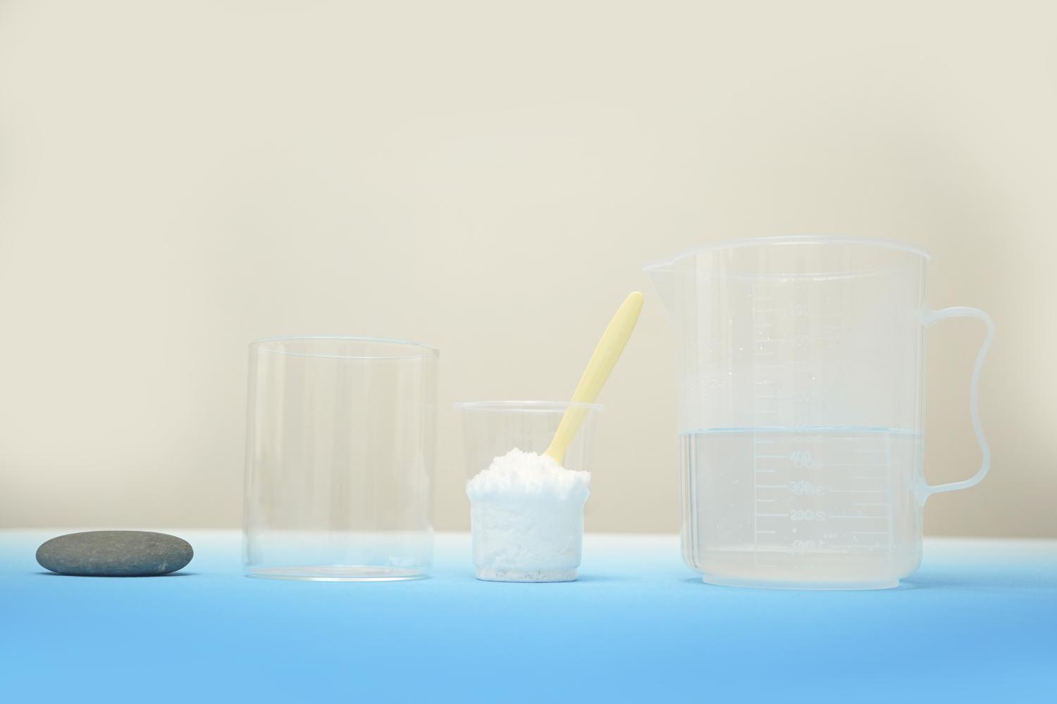 A grey stone, a clear glass, a small plastic cup of white powder, and a larger plastic jug of water are arranged on a light blue surface against a pale background.