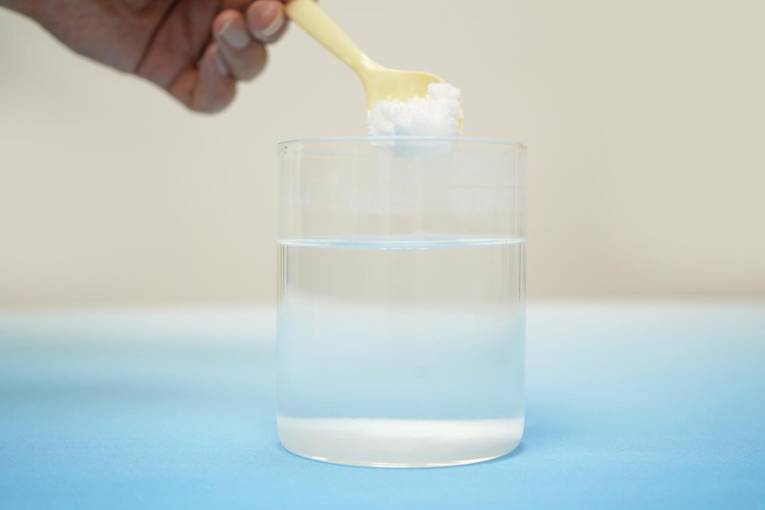 A hand adds a spoonful of white powder to a glass of water on a pale blue surface.
