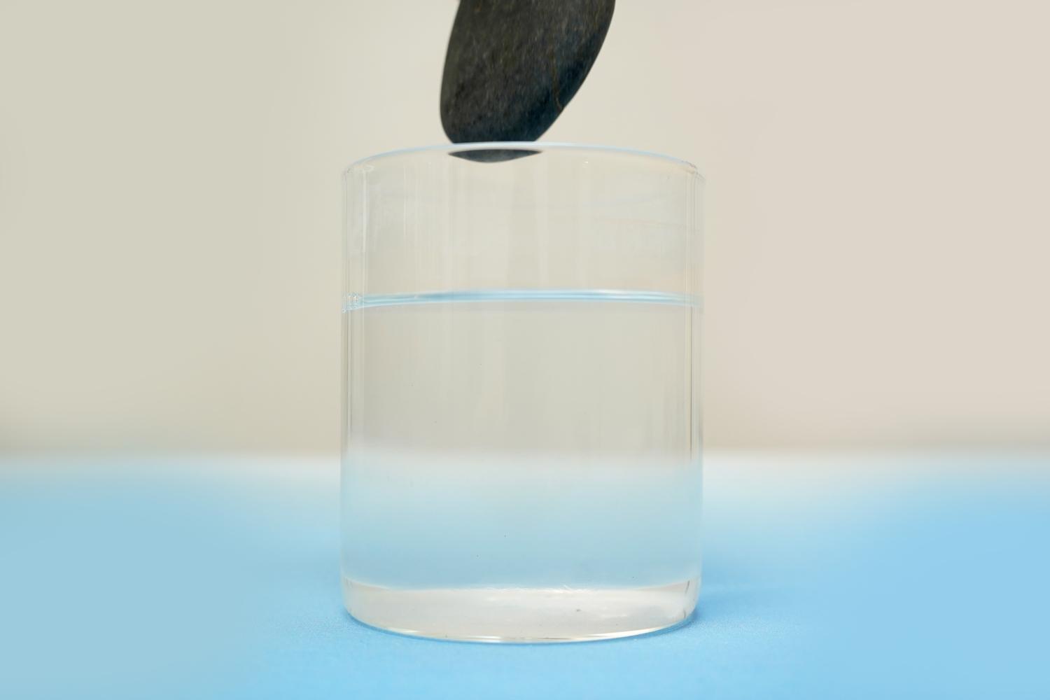 A dark grey stone is being lowered into a glass of water on a pale blue surface.